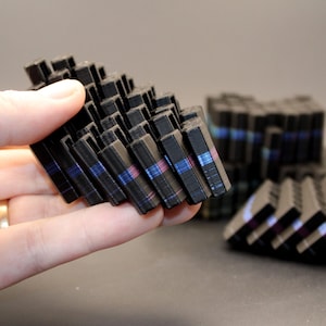 May include: A black, geometric sculpture composed of numerous small, rectangular blocks. Each block has thin stripes of blue and pink. The sculpture is held in a hand, with additional similar structures visible in the background.