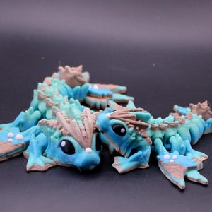 Puede incluir: Se muestran dos figuras de dragón articuladas en tonos de azul, verde azulado y marrón. Los dragones tienen escamas detalladas, cuernos y grandes ojos negros. Las figuras están posadas sobre una superficie oscura, mostrando su intrincado diseño.