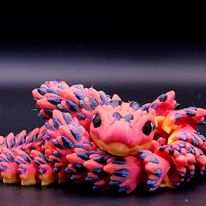 May include: A coiled snake figurine featuring a vibrant pink and yellow gradient body covered in textured, dark purple scales. The creature has large, smooth black eyes and a rounded head, with a segmented design that allows for a flexible, articulated form.