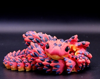 May include: A coiled articulated snake figurine featuring a vibrant pink and yellow gradient body covered in textured, dark purple scales. The creature has large, smooth black eyes and a rounded head, with a segmented design that allows for movement.
