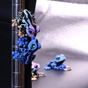 May include: A collection of colorful frog-shaped binder clips. The frogs are in shades of blue, black, purple, and cream. Some clips are attached to a metal pole, while others are lying on a dark surface. The frogs have detailed patterns and expressive eyes.
