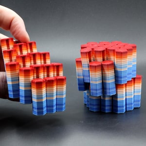 May include: A hand holding a cluster of red, orange, and blue geometric shapes. The shapes are stacked in a rectangular form. Another cluster of similar shapes is on a dark surface. The shapes have a hexagonal top.