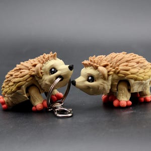 May include: Two hedgehog figurines with tan and brown quills, black eyes, and red feet. One hedgehog holds a silver keyring in its mouth. The figurines are on a dark gray surface.