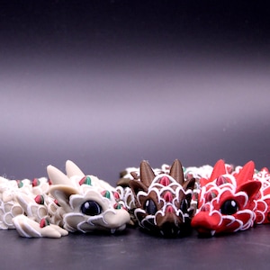 May include: Three articulated dragon figurines in white, brown, and red. Each dragon features a segmented body with white scale outlines, small green accents on the back, and large black eyes. The figures are positioned side by side.