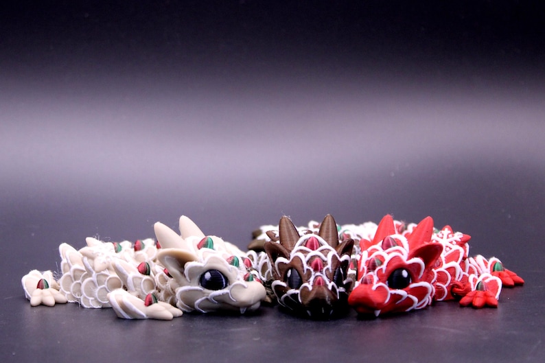 May include: Three articulated dragon figurines in white, brown, and red. Each dragon features a segmented body with white scale outlines, small green accents on the back, and large black eyes. The figures are positioned side by side.