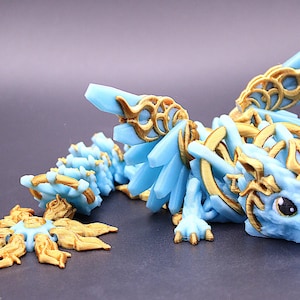 May include: A light blue dragon figurine with gold accents. The dragon has detailed wings, a spiky tail, and a stylized head. The figurine is lying down with a gold and blue decorative piece in front.