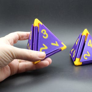 May include: Two purple and yellow triangular pyramid-shaped dice. Each face of the dice features a large yellow number. The dice are constructed with purple triangular panels held together by yellow corner connectors, creating a geometric shape.