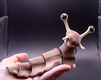 May include: A brown articulated slug figurine with a textured, segmented body and two long, upright eyestalks featuring large, round, black eyes with white highlights. The creature has a smooth, light-colored underside and a bumpy, realistic skin texture.