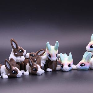 May include: A group of eight small, articulated rabbit figurines. The rabbits are in various poses and have different colour schemes. Some are brown and white, while others are a rainbow of colours.