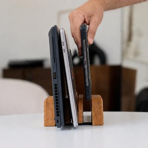 May include: A wooden stand with three vertical slots designed to hold electronic devices. The stand is crafted from light brown wood with a visible grain. Two laptops and a smartphone are positioned upright within the slots of the wooden organizer.