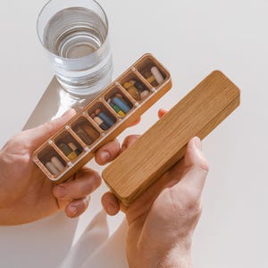 May include: A wooden pill organiser with a clear lid, filled with various colourful pills. The organiser is held open, next to a matching wooden lid. A glass of water is in the background.