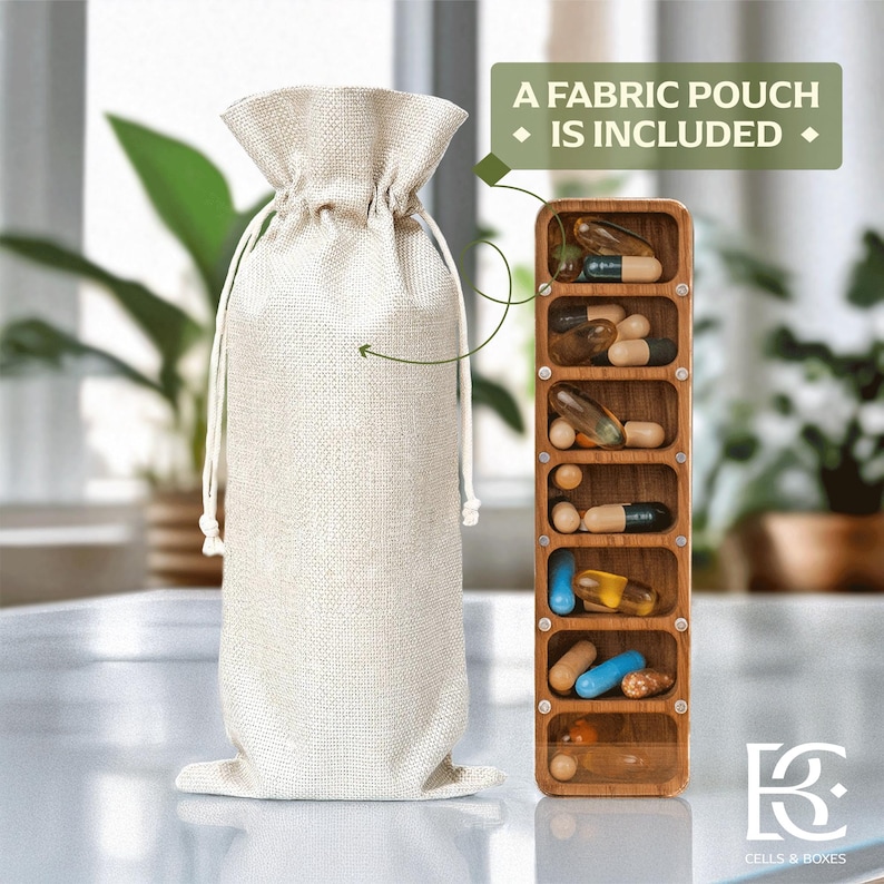 May include: A beige fabric pouch with a drawstring closure is next to a wooden pill organiser filled with various capsules and pills. The text "A Fabric Pouch Is Included" is displayed. The logo "Cells & Boxes" is in the bottom right corner.