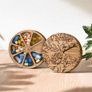 May include: A wooden pill organizer with a floral design on the lid. The organizer is divided into seven compartments, each filled with different colored pills and capsules. The lid is off to the side.