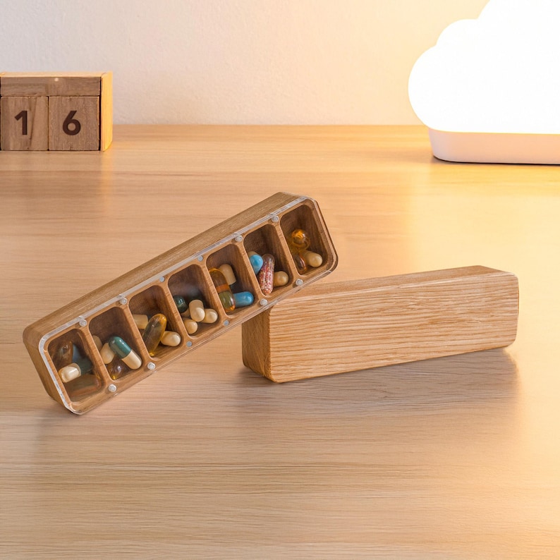 May include: A wooden pill organiser with seven compartments, each holding various colourful pills. The organiser is open, revealing the contents, and sits next to its matching lid. The wood has a natural grain.