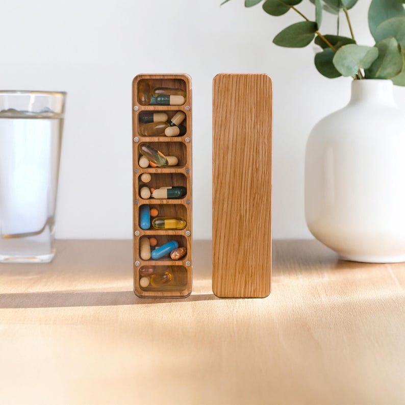 May include: A wooden pill organiser with seven compartments, each holding various pills and capsules. The organiser is open, revealing the medications inside. A separate wooden lid sits beside it. The wood has a natural grain and warm tone.