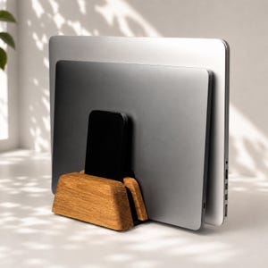 May include: A wooden laptop and smartphone stand with a natural grain finish, holding a silver laptop, a dark grey laptop, and a black smartphone in an upright, vertical position to organise multiple electronic devices on a desk surface.
