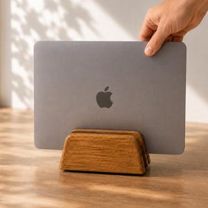 May include: A gray laptop with a centered Apple logo is positioned vertically within a rectangular wooden stand. The stand features a smooth, natural wood grain finish and a central slot designed to hold the device upright in a stable position.