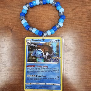 May include: A blue and silver beaded bracelet with a Blastoise Pokemon card attached. The card features the Pokemon Blastoise with the text "Vitality Spring" and "Hydro Pump".