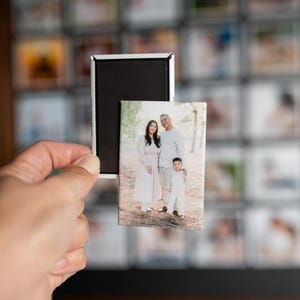 May include: A rectangular magnet with a black surface and a silver frame holds a small rectangular photo. The photo features a family of three in light-colored clothing. The background shows a wall of small, blurry photos.