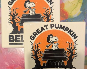 Puede incluir: Dos posavasos cuadrados de cerámica con un diseño temático de Halloween. El diseño presenta a Snoopy sentado en su caseta frente a un círculo naranja con las palabras "Great Pumpkin" arriba y "Believer" abajo. Las palabras "Since 1966" están en la parte inferior.