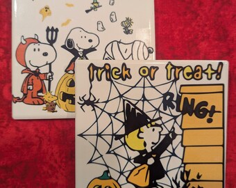 Puede incluir: Dos baldosas de cerámica cuadradas con ilustraciones de Halloween. Una baldosa presenta a Snoopy con un disfraz de diablo y otros personajes de Halloween. La segunda baldosa tiene una bruja, una calabaza y el texto "Trick or Treat!" con un diseño de telaraña.
