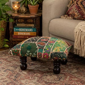 May include: A low, square ottoman featuring a patchwork fabric top with intricate floral and paisley embroidery in shades of green, red, and black. The piece is supported by four dark, rounded wooden feet and accented with brass-toned nailhead trim.
