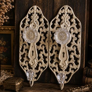 May include: Two ornate, off-white metal wall hooks with clear glass knobs and keyhole details. The hooks feature an intricate, scrolling design. The background includes a candle, dried flowers, and antique decor.