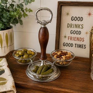 May include: A three-section serving tray featuring a central dark wood handle with a silver-toned metal ring at the top. The tray includes three clear glass bowls arranged around the base, currently holding green olives, mixed nuts, and pickles.
