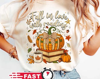 May include: Cream-colored t-shirt featuring a fall-themed graphic. The design includes a pumpkin with a plaid bow, books, a coffee mug with a maple leaf, and the text "Fall in love with reading". Autumn leaves and acorns complete the design.