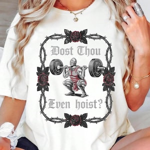 May include: Cream-colored t-shirt featuring a weightlifting knight graphic within a thorny rose frame. The text "Dost Thou Even hoist?" is printed above and below the knight. The design includes black and red accents.