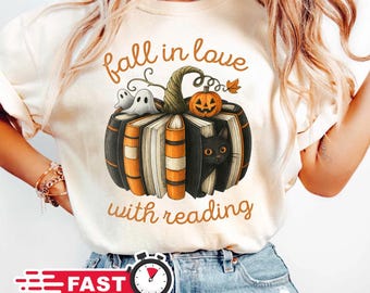 Peut inclure: T-shirt crème avec un motif d'Halloween. Le design comprend une citrouille faite de livres, un chat noir et le texte "fall in love with reading". Un autocollant de livraison rapide est visible.