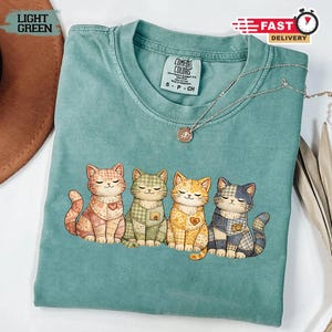 Può includere: Una maglietta Comfort Colors verde chiaro con quattro gatti dei cartoni animati con motivi patchwork. Ogni gatto ha un cuore sul petto. È visibile una collana d'argento con un ciondolo. La maglietta ha uno scollo tondo.