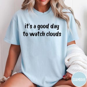 May include: A light blue short-sleeved t-shirt featuring the black text "it's a good day to watch clouds" printed across the chest. The garment has a crew neckline and a relaxed fit, with a white textured fabric item held near the side.
