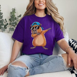 May include: A purple t-shirt featuring a cartoon capybara wearing sunglasses and a blue cap, dabbing with its arm. The capybara is brown and tan. The shirt is a solid color and has a crew neck.