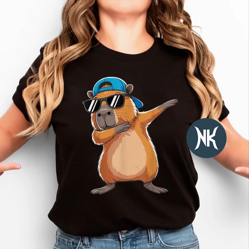 May include: Black t-shirt featuring a cartoon capybara wearing sunglasses and a blue baseball cap, striking a dabbing pose. The capybara is tan with a gray nose and feet. The shirt has a round neckline.