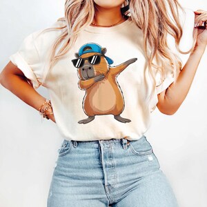 May include: Beige t-shirt featuring a cartoon capybara wearing sunglasses and a blue cap, striking a dab pose. The capybara is brown with a gray nose and feet. The shirt has rolled-up sleeves and is paired with blue jeans.