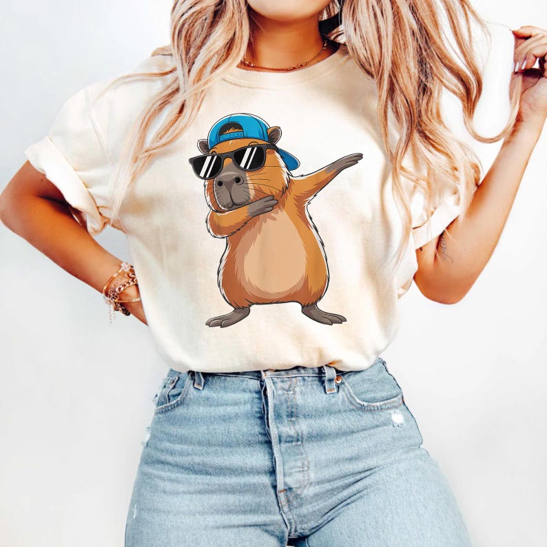 May include: Beige t-shirt featuring a cartoon capybara wearing sunglasses and a blue cap, striking a dab pose. The capybara is brown with a gray nose and feet. The shirt has rolled-up sleeves and is paired with blue jeans.