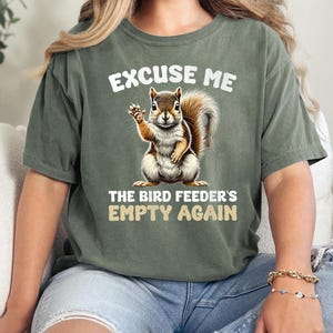 Op de afbeelding: Een groen T-shirt met een witte afbeelding van een eekhoorn die een bord vasthoudt met de tekst: "Excuse me, the bird feeder's empty again."