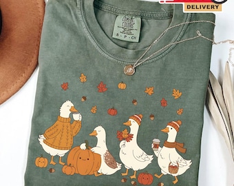 Puede incluir: Camiseta verde musgo Comfort Colors con un diseño otoñal. El gráfico muestra cuatro patos, calabazas y hojas de otoño. Un pato lleva un suéter, otro un gorro y bufanda, y otros sostienen tazas de café y hojas. También se ve un collar plateado.