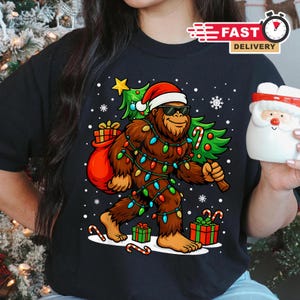 May include: Black t-shirt featuring a cartoon Bigfoot wearing a Santa hat and sunglasses, carrying a Christmas tree and a sack of gifts. The Bigfoot is wrapped in Christmas lights, with snowflakes and candy canes around it.