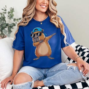 May include: A blue t-shirt featuring a cartoon capybara wearing sunglasses and a blue cap, posed in a dabbing stance. The capybara is brown and tan, with a white outline. The shirt is a solid blue color.