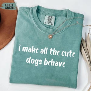May include: A light green short-sleeved t-shirt with a crew neckline and a Comfort Colors label at the inner collar. The front features the white text "i make all the cute dogs behave" in a rounded font. A thin gold chain necklace rests on the fabric.