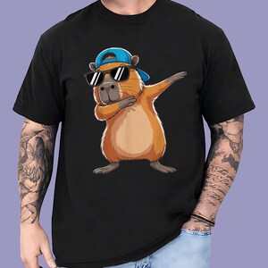 May include: Black t-shirt featuring a cartoon capybara wearing sunglasses and a blue baseball cap, with its arm raised in a dabbing pose. The capybara is brown and tan, with a gray nose and black sunglasses.