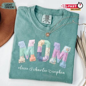 Może przedstawiać: Jasnozielona koszulka Comfort Colors z napisem "MOM" naszytym z kolorowej tkaniny patchworkowej. Poniżej wydrukowano imiona "elisie", "charlie" i "sophia". Srebrny naszyjnik z wisiorkiem jest zarzucony na koszulkę.