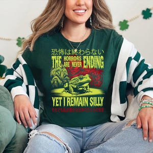 May include: A green long-sleeved shirt with a frog graphic and the text "The Horrors Are Never Ending Yet I Remain Silly" in yellow and green. The text is in Japanese and English.