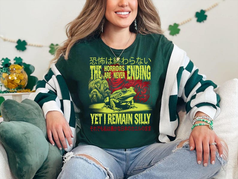 May include: A green long-sleeved shirt with a frog graphic and the text "The Horrors Are Never Ending Yet I Remain Silly" in yellow and green. The text is in Japanese and English.