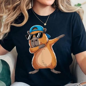 May include: Black t-shirt featuring a cartoon capybara wearing a blue baseball cap and sunglasses, dabbing with its arm. The capybara is brown and tan with a light tan belly. The shirt is a dark navy blue.