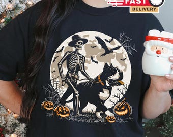 May include: Black t-shirt featuring a Halloween design. A skeleton in a hat walks a large black and white dog in front of a full moon, with bats, spiderwebs, and pumpkins. The shirt has a "Fast Delivery" logo.