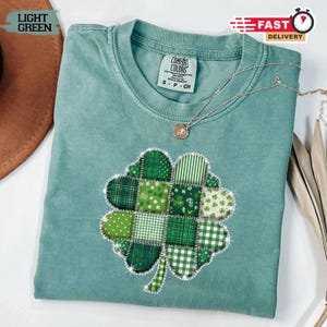Può includere: T-shirt Comfort Colors verde chiaro con un design a trifoglio patchwork. Il trifoglio è composto da vari motivi e trame verdi, delineati con abbellimenti scintillanti. Una collana d'argento con un ciondolo.