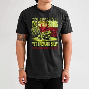 May include: Black t-shirt with a green and red graphic of a frog and the text "The horrors are never ending, yet I remain silly". The text is in Japanese and English.